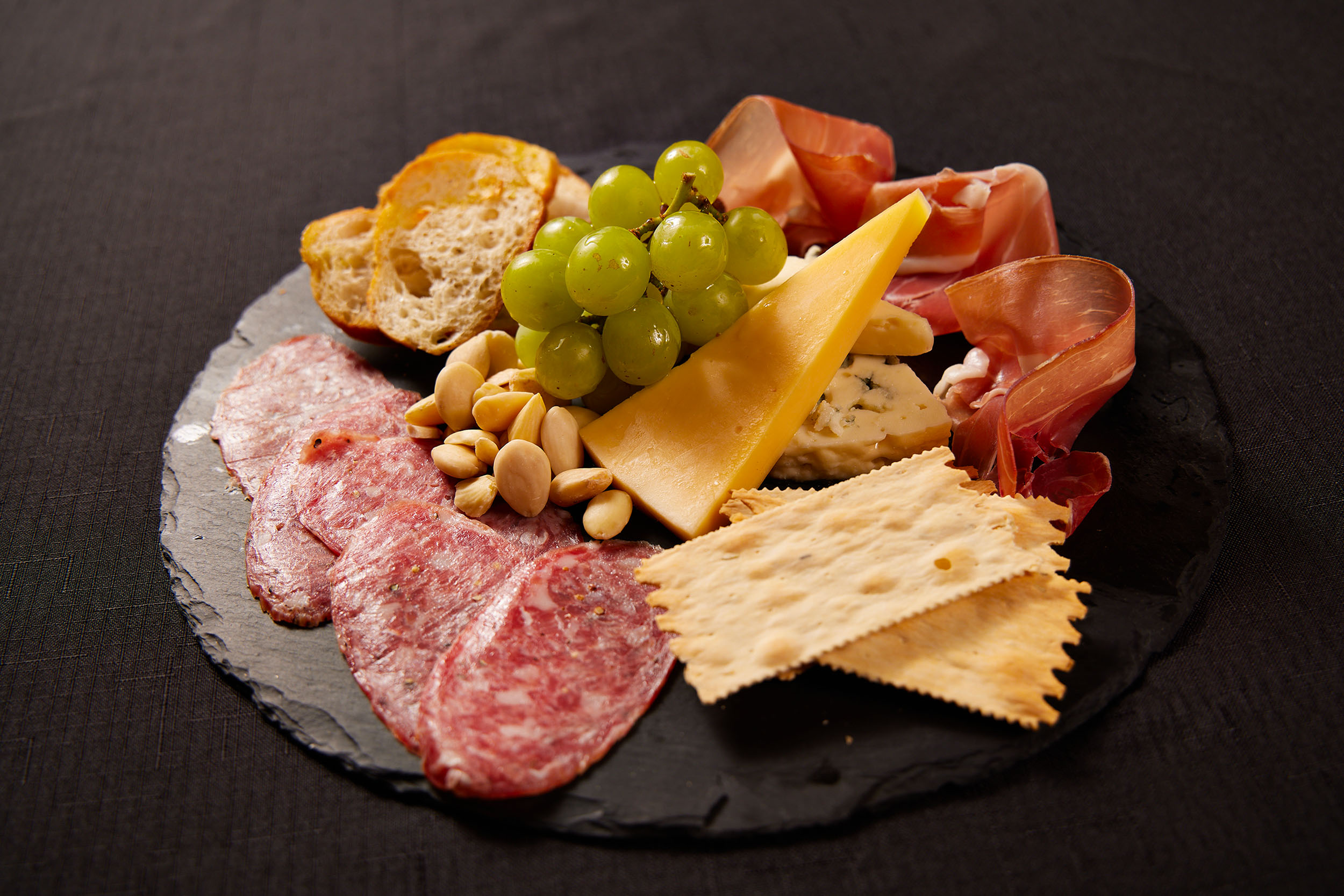 A charcuterie plate on a dark slate includes salami, prosciutto, cheese slices, blue cheese, crackers, bread slices, green grapes, and almonds—creating a world-class dining experience—arranged neatly on a black tablecloth.