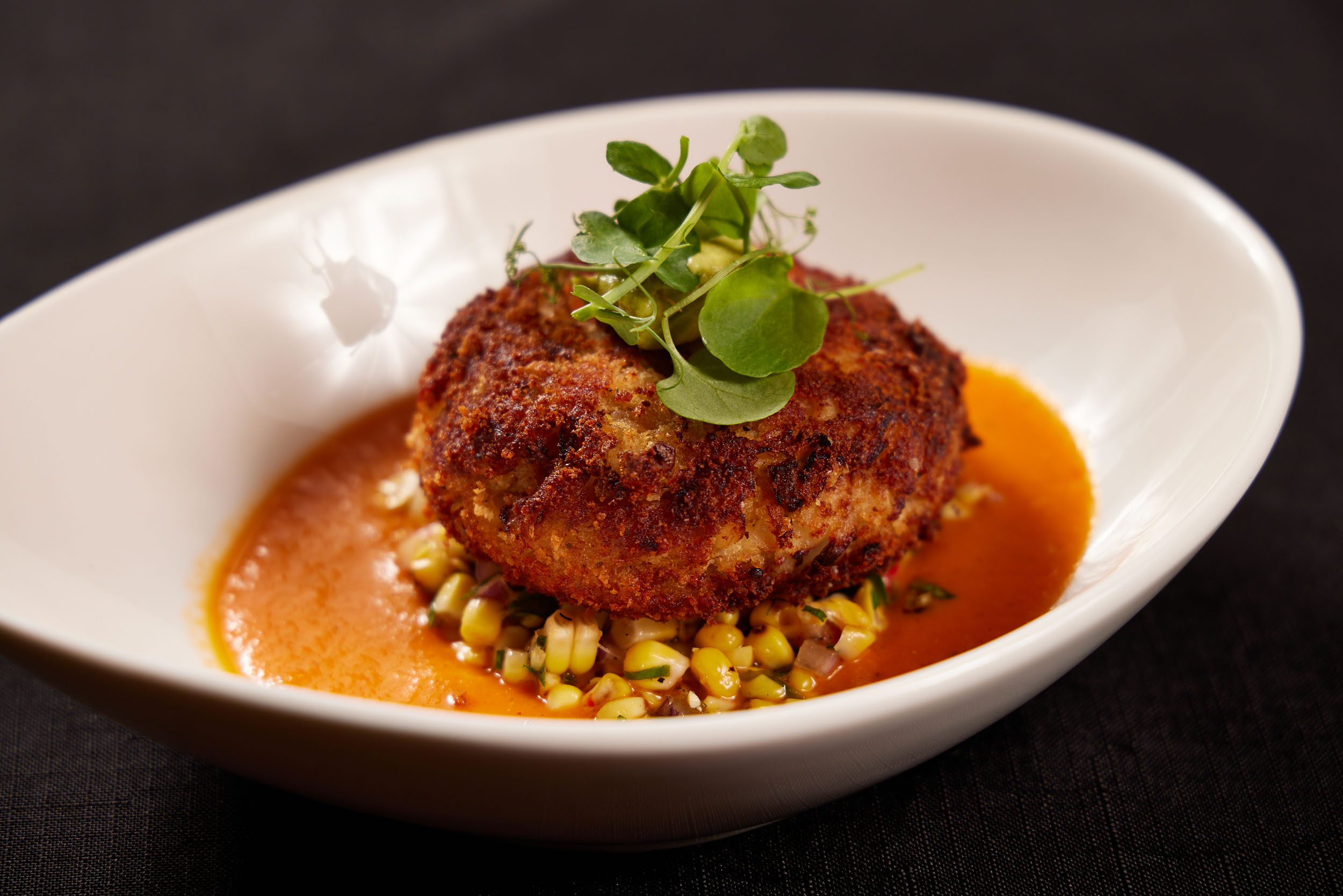 A golden-brown crab cake sits atop a bed of corn relish in a white bowl, surrounded by an orange sauce. This world-class dining dish is garnished with fresh green microgreens, and the dark background highlights the plated food.