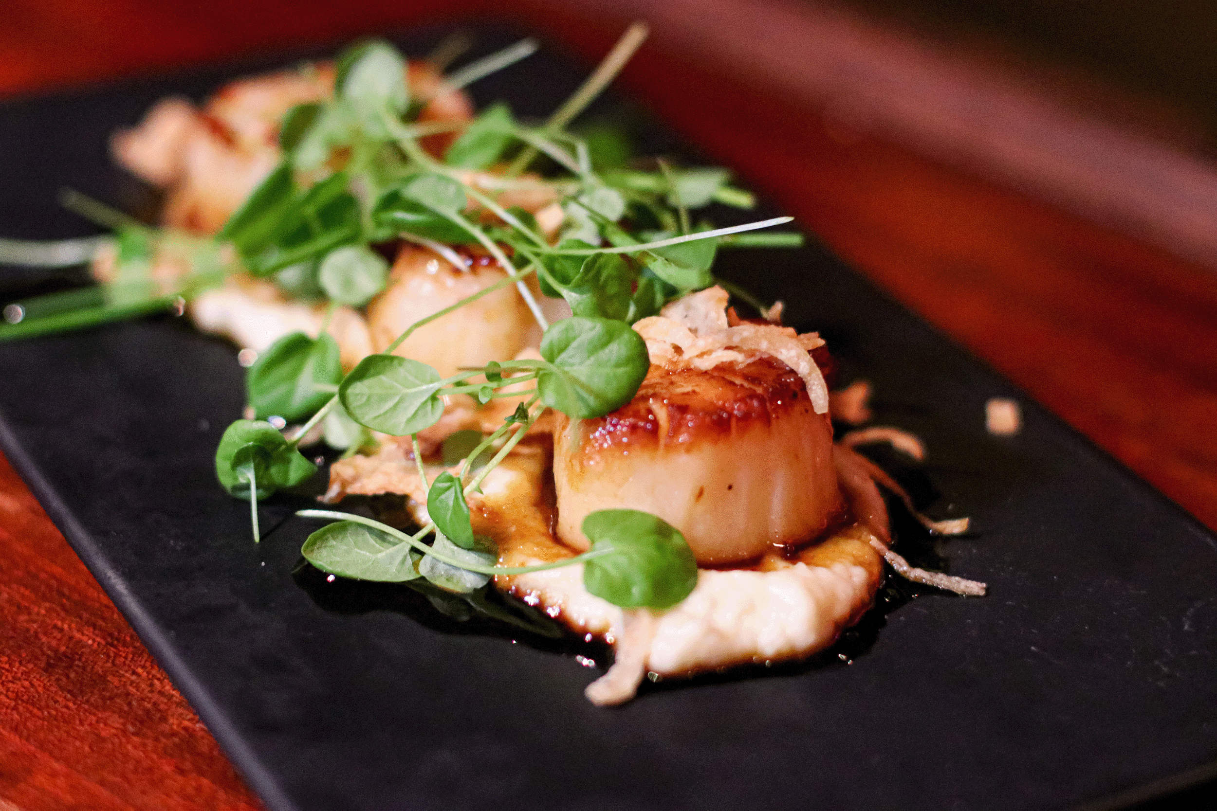 Seared scallops are plated on a black rectangular dish, topped with microgreens and crispy shallots, resting on a creamy sauce. The elegant presentation and fresh flavors embody the essence of world-class dining against the wooden table backdrop.