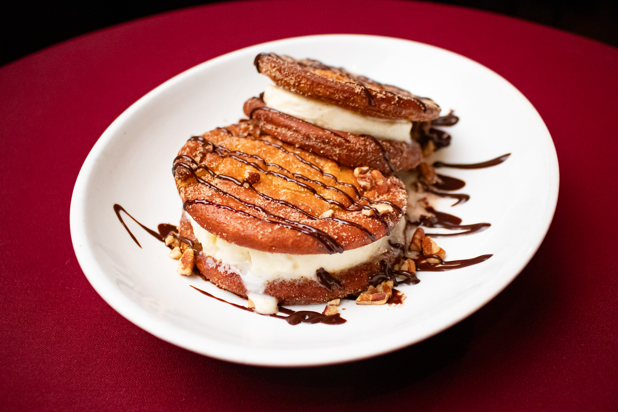 A white plate holds two ice cream sandwiches made with golden brown, sugar-dusted churros and vanilla ice cream—a world-class dining treat—drizzled with chocolate sauce and sprinkled with chopped nuts, set against a deep red tablecloth.