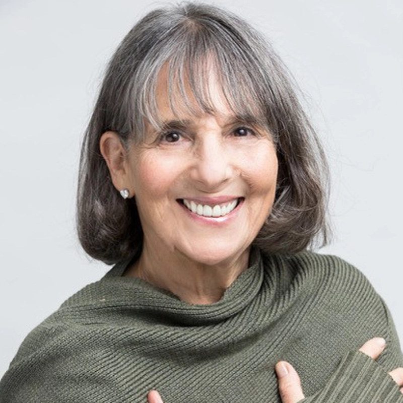A smiling older woman, Loni Ackerman Kennedy, with gray, shoulder-length hair and bangs wears a textured olive green wrap and pearl earrings, posing against a light gray background. She looks directly at the camera with her arms crossed gently over her chest.