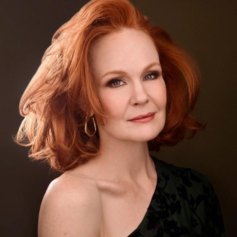 A woman with fair skin and voluminous red hair, reminiscent of Kate Baldwin, poses against a dark background. She wears a black, one-shoulder top and gold hoop earrings, looking confidently at the camera with a slight smile and soft makeup.