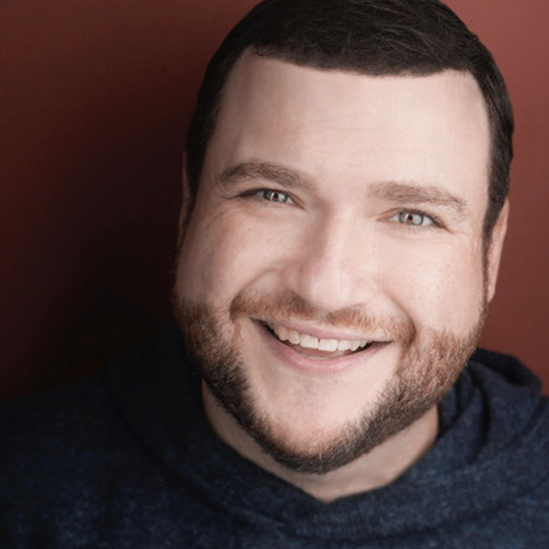 A smiling man with short dark hair and a trimmed beard—Todd Buonopane—is wearing a navy blue hoodie. He faces the camera, bright-eyed and friendly, posed in front of a solid reddish-brown background.