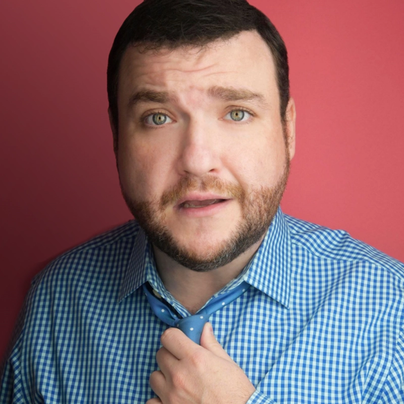 A man with short brown hair, a beard, and green eyes looks at the camera with a slightly concerned expression. Reminiscent of Todd Buonopane, he wears a blue checked shirt and loosely holds a light blue polka dot tie against a pinkish-red background.