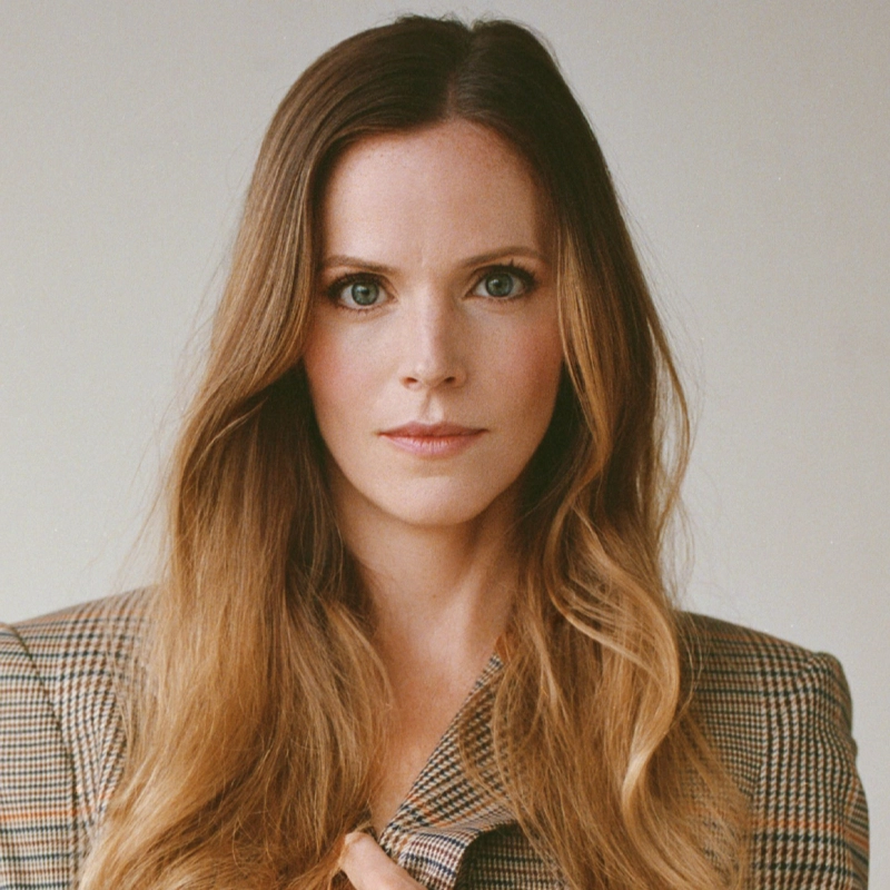 Katie Rose Clarke, a woman with long, wavy light brown hair and green eyes, stands against a plain light background in a brown plaid blazer, looking directly at the camera with a neutral expression.