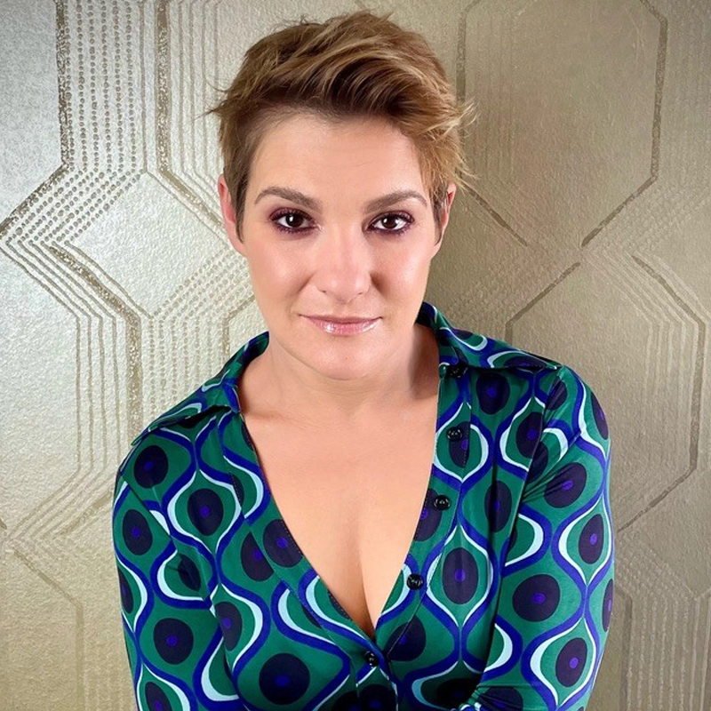A person with short, light brown hair and light makeup smiles softly at the camera. They wear a green and blue patterned shirt with a deep neckline. The background features a gold geometric wallpaper with an Art Deco design.