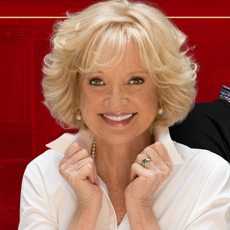 A smiling older woman, resembling Christine Ebersole, with short, curly blonde hair, wears a white collared shirt and pearl earrings. She holds her collar with both hands and stands against a red background, with only part of another person visible beside her.