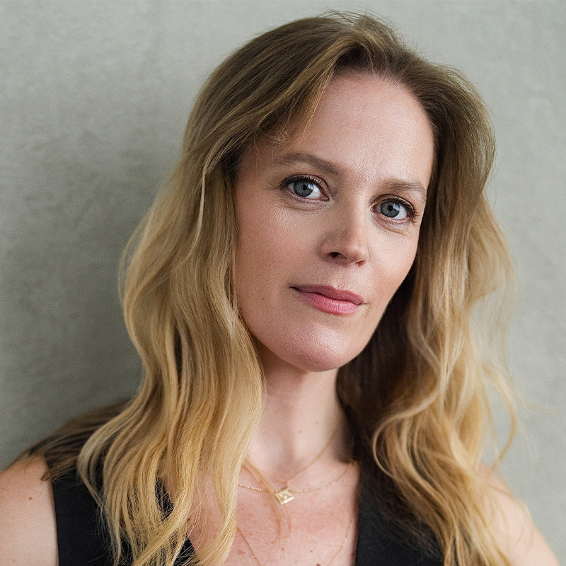 Actress Elizabeth Stanley, a woman with wavy blonde hair and blue eyes, looks directly at the camera while wearing a dark sleeveless top and a delicate gold necklace with a square pendant.