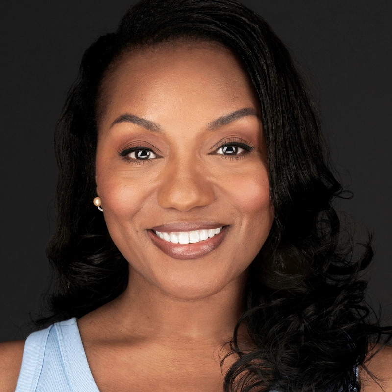 A woman with medium-dark skin, long wavy dark hair, and brown eyes smiles warmly at the camera. She wears a light blue sleeveless top, natural makeup, and small gold stud earrings. The background is plain black.