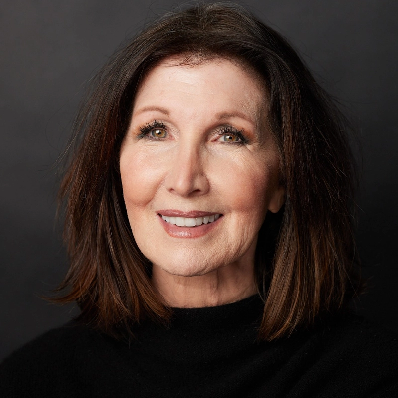 A headshot of actress Joanna Gleason, a smiling middle-aged woman with shoulder-length brown hair and warm-toned makeup. She is wearing a black top against a solid dark grey background.