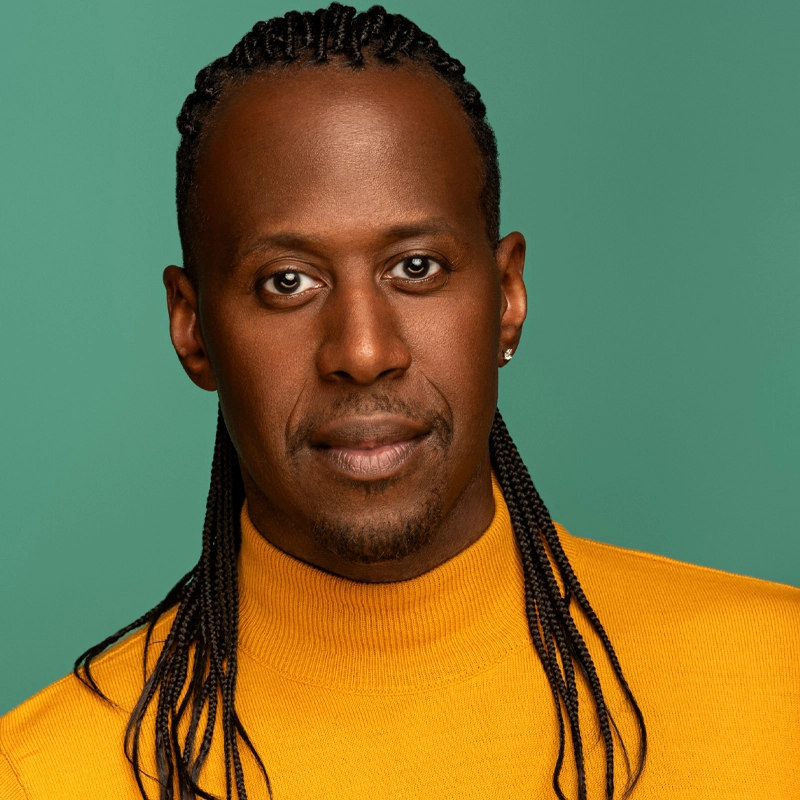 Marcus Paul James, a man with braided hair, a trimmed beard, and brown skin, wears a yellow turtleneck sweater. He has a stud earring in his left ear and looks directly at the camera with a calm expression against a solid teal background.