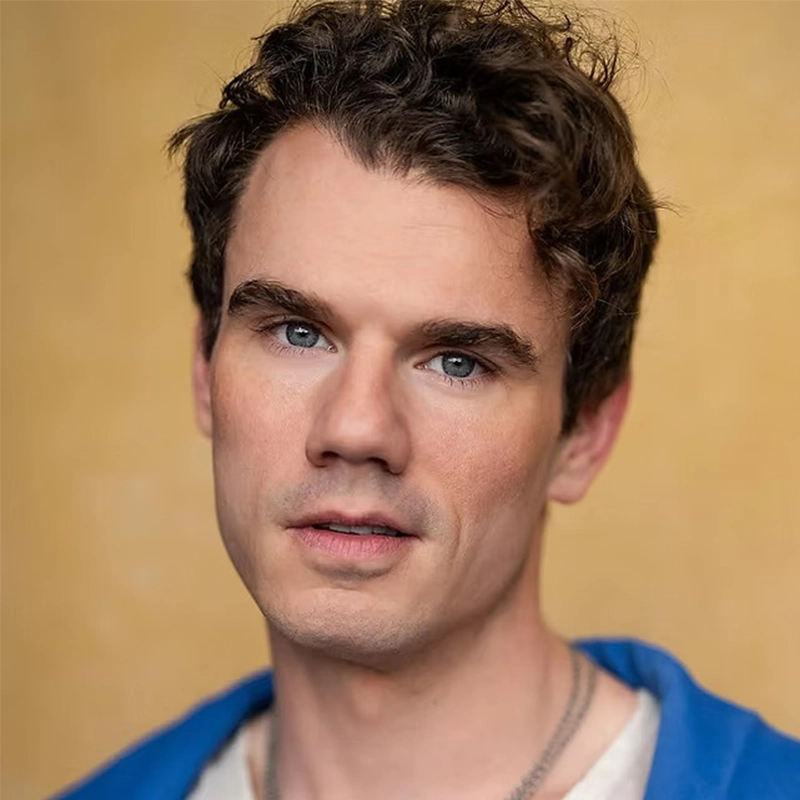 Jay Armstrong Johnson, a young man with short, wavy brown hair and blue eyes, gazes at the camera. He has fair skin, light stubble, and wears a blue jacket over a gray shirt against a soft, warm beige studio background.