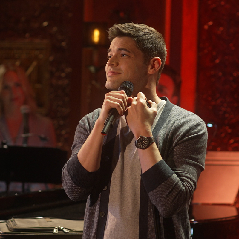 Jeremy Jordan stands on stage holding a microphone near his chin, wearing a gray cardigan over a light shirt and a wristwatch. He looks thoughtful, with warm stage lights and a blurred background featuring a piano and another person.