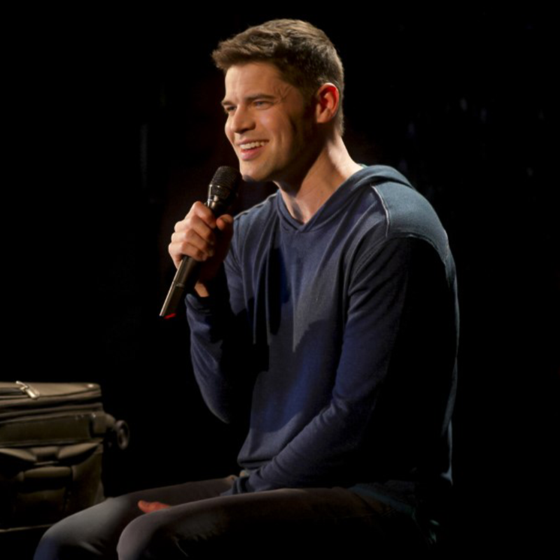 Jeremy Jordan, with short brown hair, sits on a stool, smiling and holding a microphone in his right hand. He wears a navy blue long-sleeve hoodie and dark pants. The stage lighting highlights his face against the dark background and small suitcase.