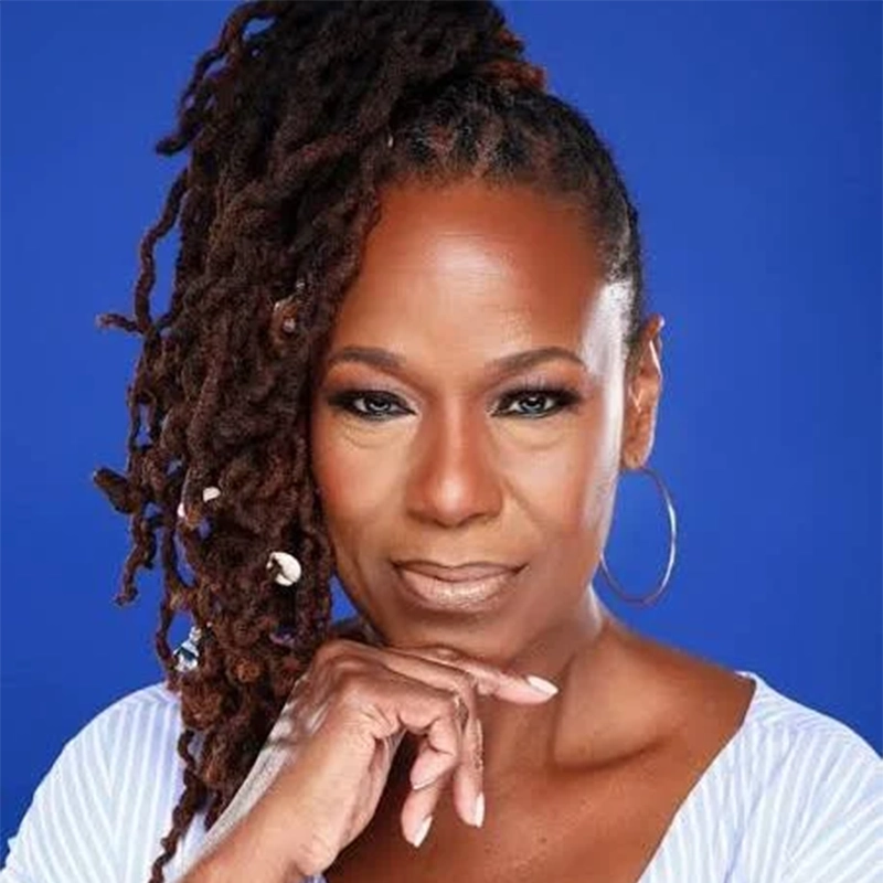 A confident Black woman with long, styled locs pulled up to one side poses against a blue background. She wears hoop earrings, subtle makeup, and a light top, resting her chin on her hand while looking directly at the camera.