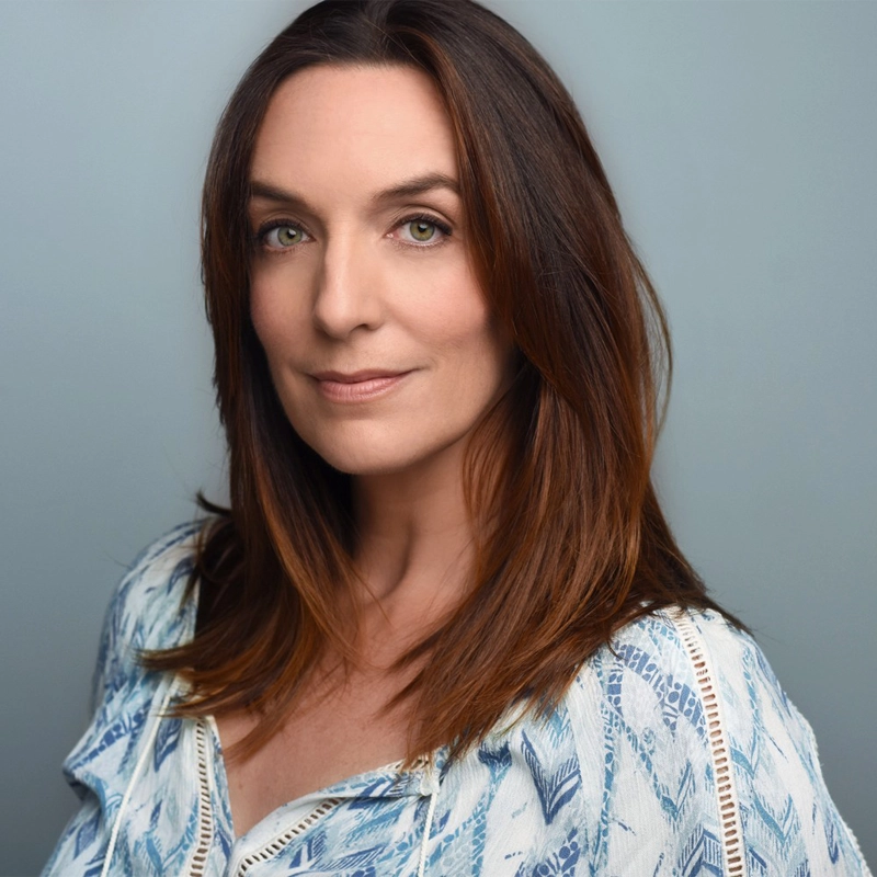 Julia Murney, with long, straight brown hair and light green eyes, looks calmly at the camera. She wears a patterned white and blue blouse and stands against a simple, light grey background, her expression neutral and confident.