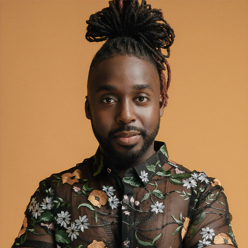 Actor and singer Mykal Kilgore wears a black, sheer, floral-embroidered button-down shirt. With his dreadlocks styled in a high bun and a full beard, he looks directly at the camera against a solid tan background.