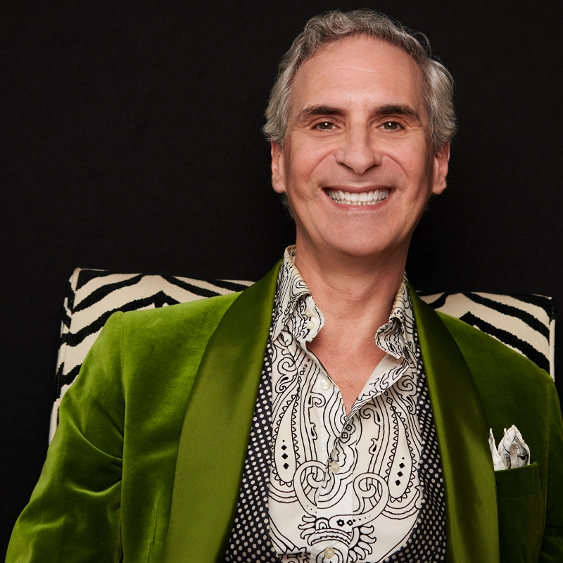 A smiling man with gray hair sits against a zebra-patterned background. He wears a green velvet blazer over a black and white patterned shirt and vest, with a white pocket square visible. The image is brightly lit, highlighting his cheerful expression.
