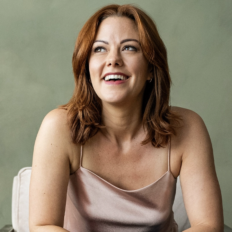 Kate Rockwell, with shoulder-length auburn hair, smiles joyfully while looking slightly to her left. She wears a light pink satin camisole and sits against a soft green background, her expression relaxed and radiant.