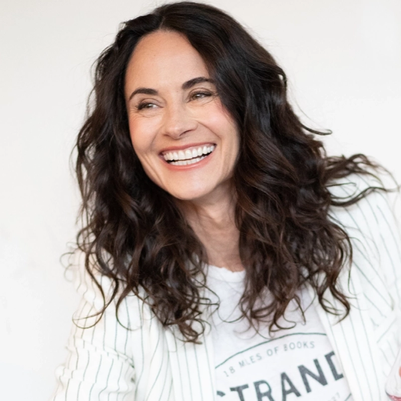 A woman with long, wavy dark hair smiles brightly. She wears a white shirt with a circular logo and a white blazer with thin stripes. The background is plain and light, making her stand out. She looks happy and relaxed, facing slightly to the right.