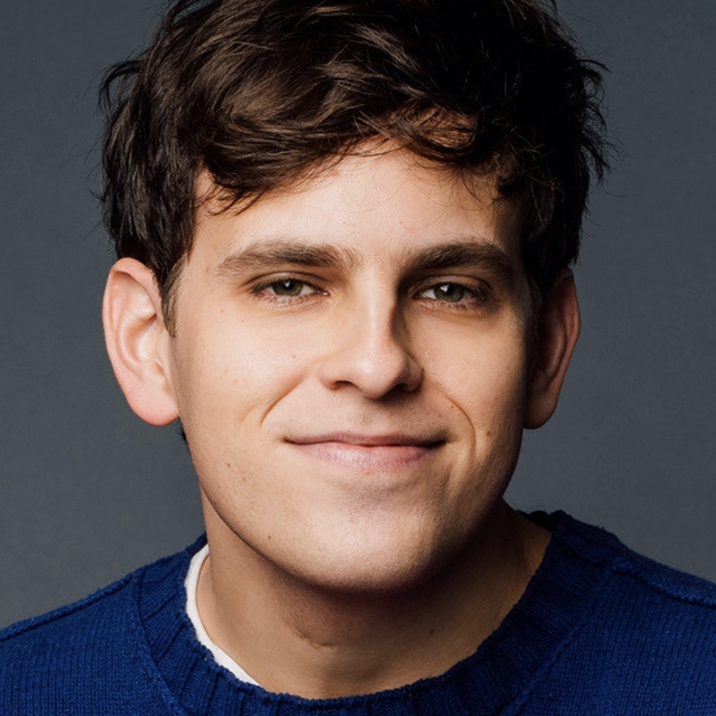 A young man with short brown hair, resembling Taylor Trensch, smiles gently at the camera. He is wearing a dark blue knit sweater over a white shirt, with a plain dark background highlighting his face and casual, friendly expression.