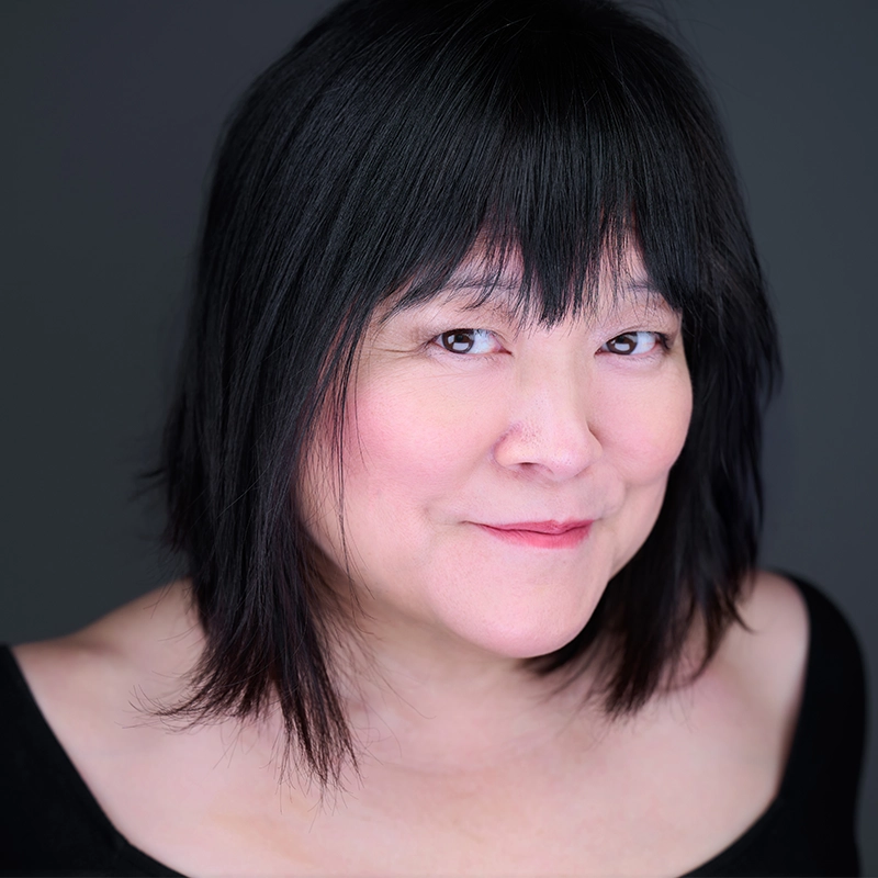 Natascia Diaz, with straight, shoulder-length black hair and bangs, smiles softly at the camera. She has fair skin with a subtle blush and wears a black off-the-shoulder top. The dark gray background adds to the gentle, professional portrait vibe.