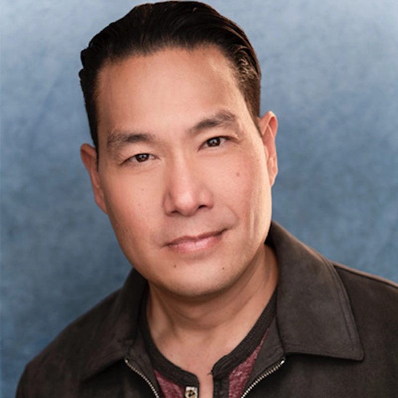 A man with short, dark hair combed back is wearing a dark jacket over a maroon shirt. He faces the camera with a slight smile against a soft, blue gradient background. The even lighting subtly evokes the style of Natascia Diaz portraits.