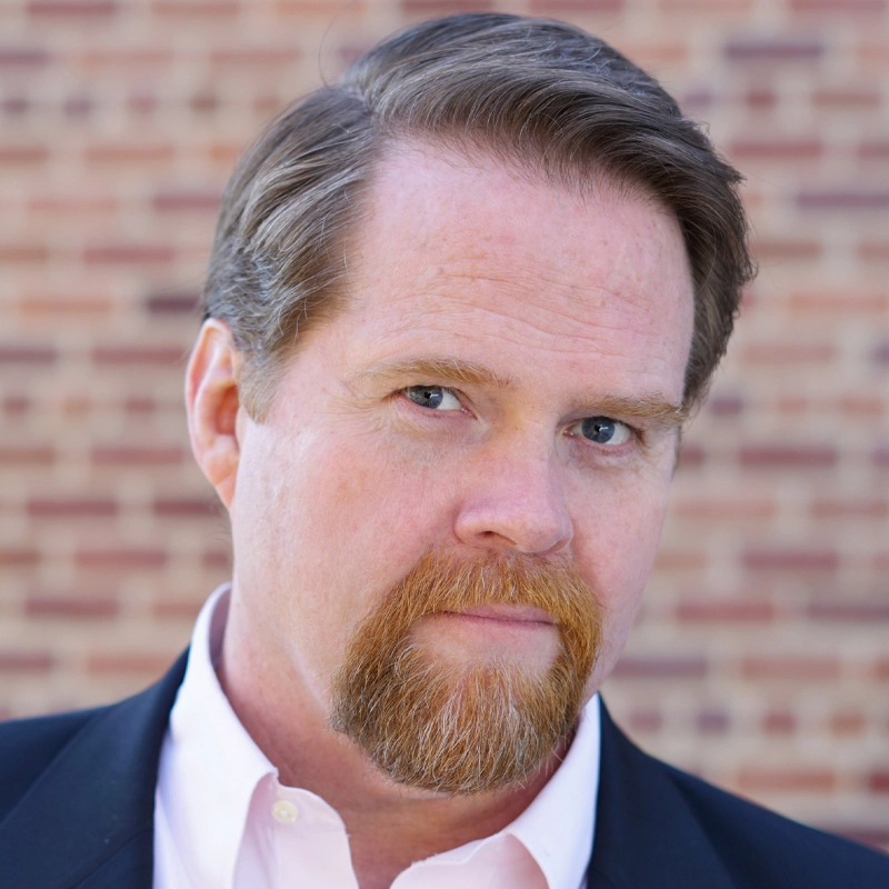 John Treacy Egan, a man with light skin, reddish-brown hair, and a matching goatee, looks seriously at the camera. He wears a white dress shirt and dark blazer. The background is out of focus, showing a red brick wall.