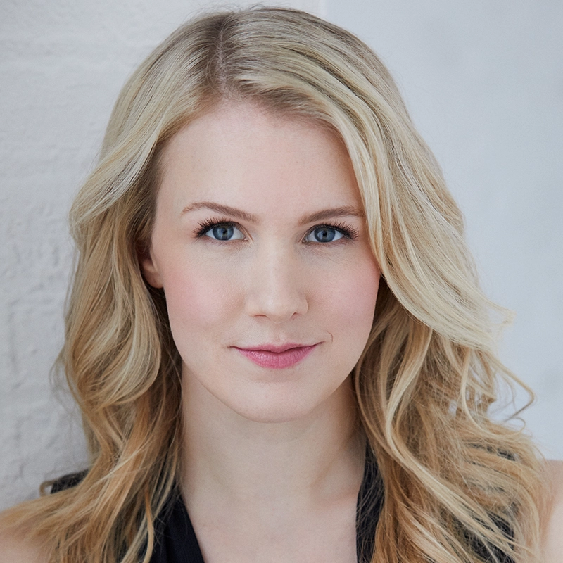 A young woman with long, wavy blonde hair and blue eyes faces the camera, slightly smiling. Reminiscent of Ginna Claire Mason, she is wearing a sleeveless dark top and light makeup. The background is softly lit with a white textured wall on the left.
