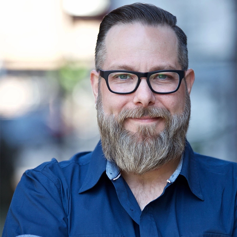 Evan Harrington, a man with light skin, a full brown and gray beard, and short hair styled back, wearing black glasses and a blue collared shirt, smiles gently at the camera against a softly blurred outdoor background.