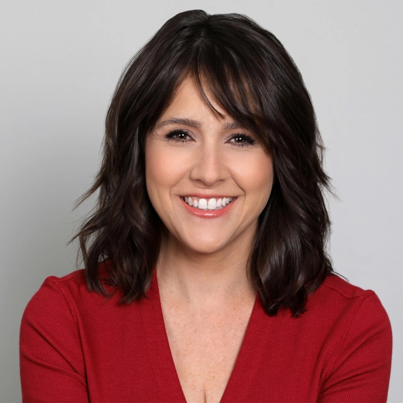 A woman with medium-length wavy dark brown hair and side-swept bangs smiles at the camera. She is wearing a red V-neck top and has a light skin tone. The background is plain light gray, giving a professional, approachable feel.