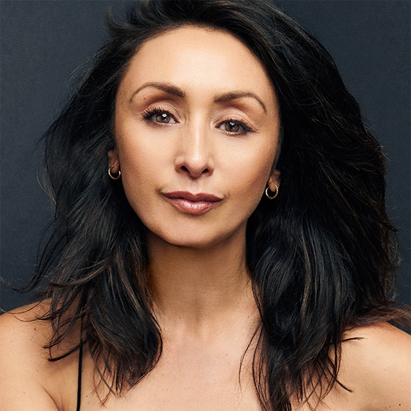Natascia Diaz, with long, wavy dark hair and light brown skin, stares confidently at the camera. She wears small hoop earrings and a black strappy top against a dark background, her calm expression enhanced by subtle makeup.