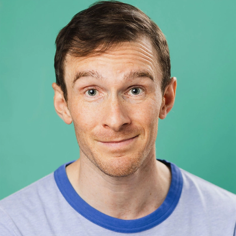 A man with light skin and short brown hair smiles slightly. He is wearing a light purple T-shirt with blue trim and is posed against a plain teal background. He has a friendly, approachable expression and is looking directly at the camera.