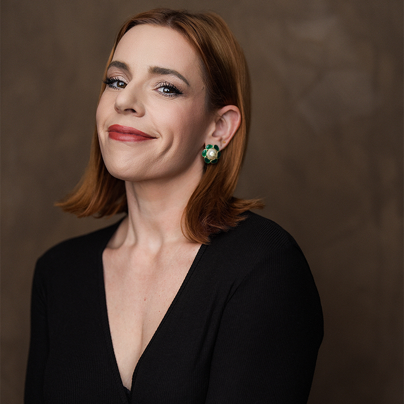 A portrait of actress and singer Alysha Umphress with shoulder-length auburn hair, wearing a black V-neck top. She has a subtle smile and gold flower-shaped earrings with green enamel petals and a pearl center.