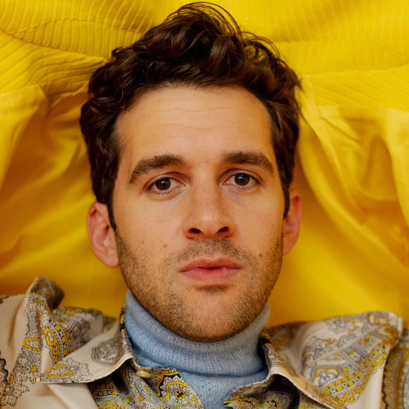 Adam Chanler-Berat, a young man with short, curly brown hair and light facial hair, looks directly at the camera. He wears a light blue turtleneck and a patterned shirt against a vivid yellow background and fabric, creating a striking contrast.