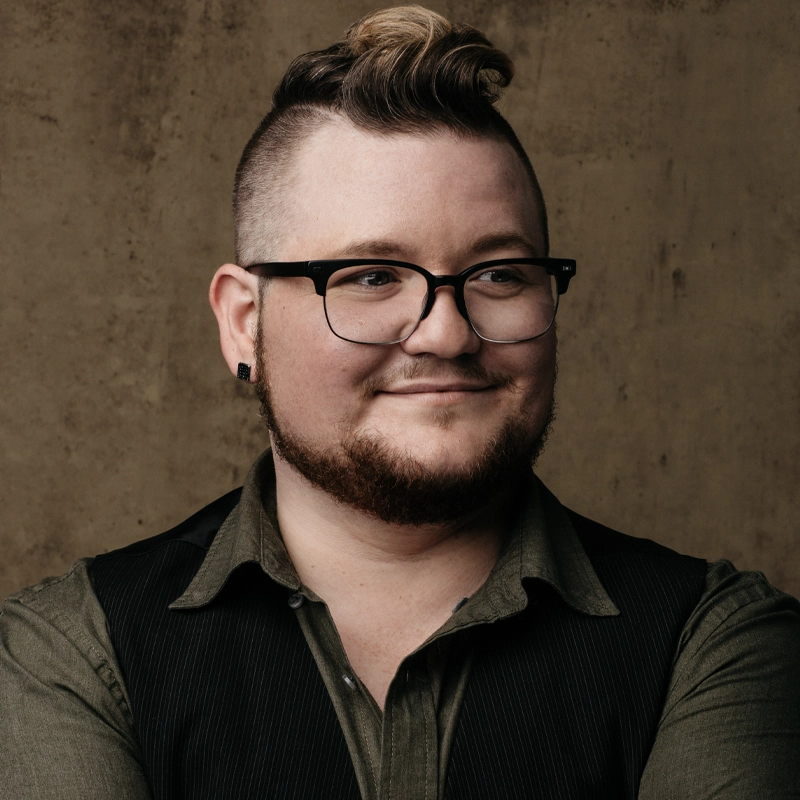 A person with short hair on the sides and longer, wavy hair on top poses against a brown background. Donnie Cianciotto smiles softly, wearing glasses, black earrings, an olive shirt, and a black vest, looking slightly to the side.