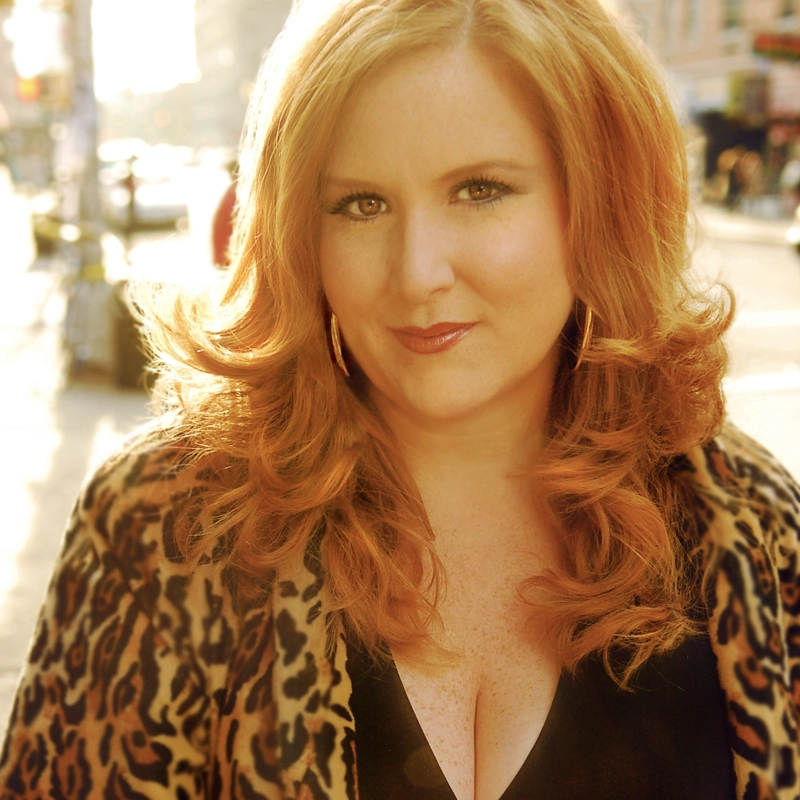 A woman with long, wavy red hair and light skin stands outdoors in sunlight. She wears a leopard print jacket over a black top and large hoop earrings. She looks directly at the camera with a confident expression, while a city street scene is blurred in the background.