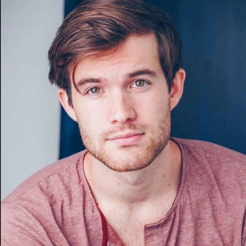 Brandon Kalm, a young man with short brown hair and light stubble, gazes at the camera with a slight smile. He wears a mauve scoop-neck shirt and stands before a striking two-tone background of white and dark blue.