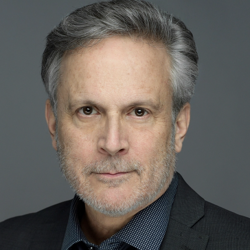 A middle-aged man with gray hair and a short beard wearing a dark blazer over a dotted shirt. He has a neutral expression and is looking directly at the camera. The background is plain gray.
