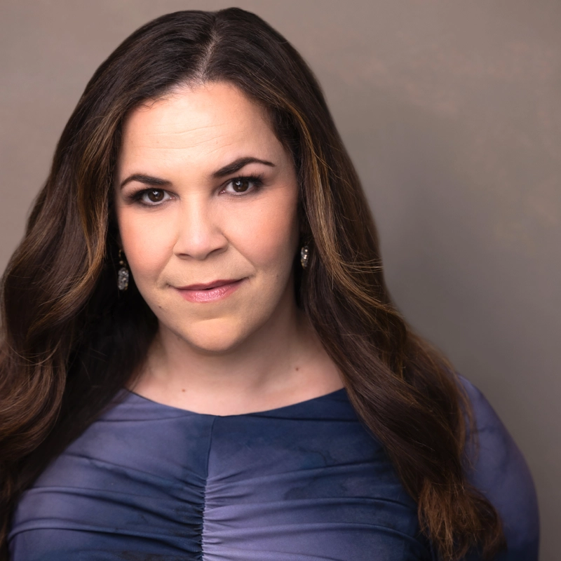 A woman with long, wavy brown hair and fair skin gazes confidently at the camera. Reminiscent of Lindsay Mendez, she wears a dark blue, ruched top and dangling earrings against a neutral background that highlights her composed expression.