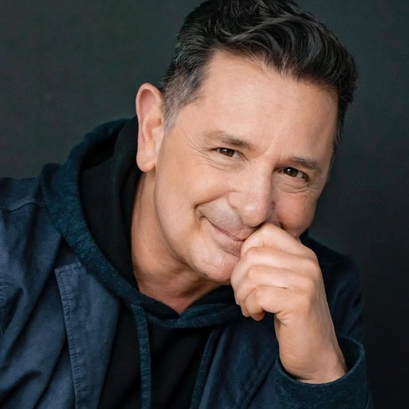 A middle-aged man with short dark hair smiles gently at the camera, resting his chin on his hand. He is wearing a dark hoodie and a navy jacket. The background is plain and dark, highlighting his expressive, friendly face.