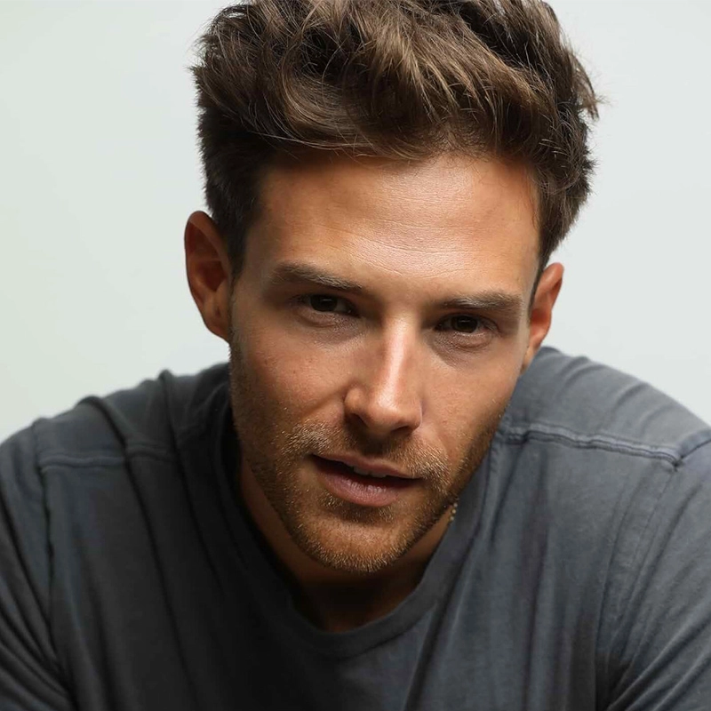A man with short, tousled brown hair and trimmed facial stubble—resembling Ben Rappaport—looks at the camera. He wears a grey crew-neck t-shirt and has a slight, thoughtful expression against a plain, softly lit background.
