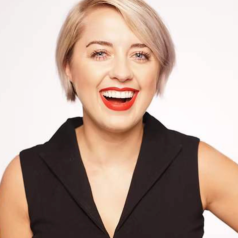 Mollie Craven, with short blonde hair, smiles broadly, showing teeth and bright red lipstick. Dressed in a sleeveless black collared top against a plain white background, she exudes confidence and happiness.