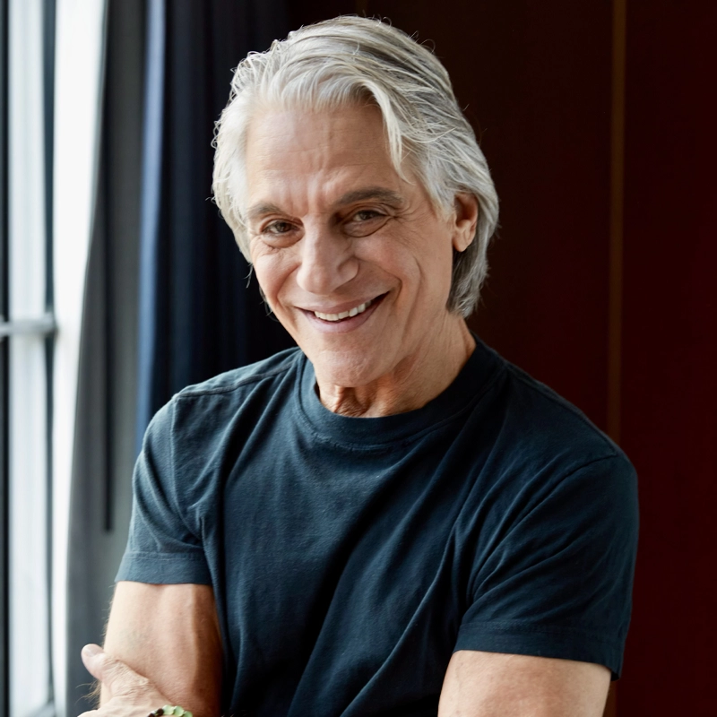 An older man with white hair, wearing a dark short-sleeve t-shirt, stands indoors by a window with his arms crossed and smiles warmly at the camera. The background features dark curtains and a softly lit wall.