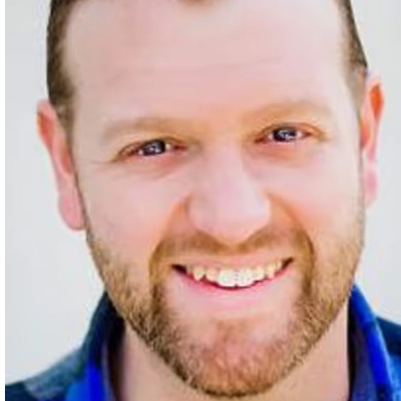 A close-up photo of a smiling man with light skin, short brown hair, and a neatly trimmed beard. He is wearing a blue shirt and is facing the camera. The background is plain and out of focus.