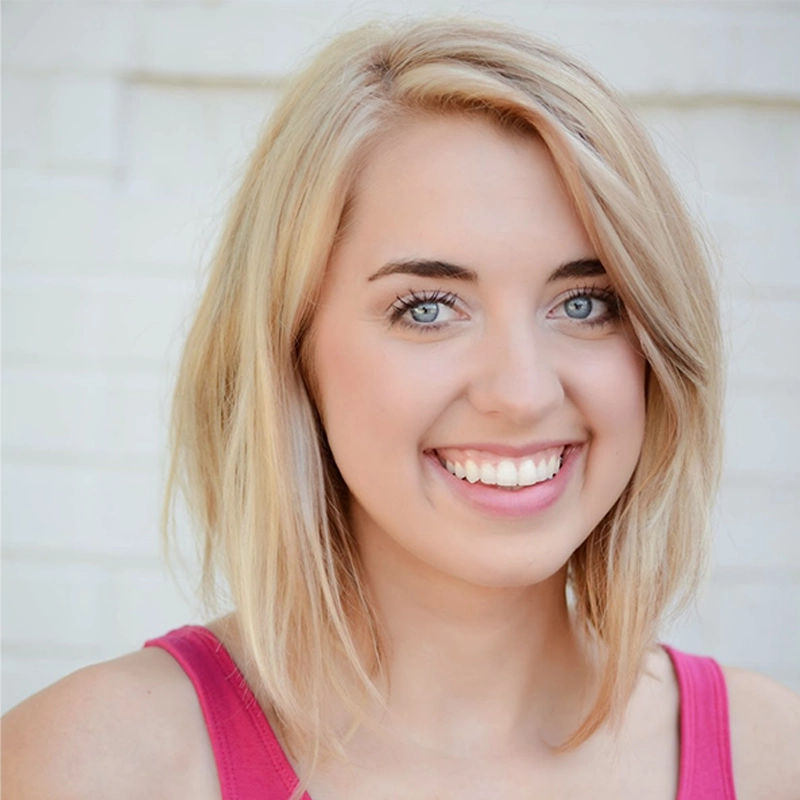 Mollie Craven, a young woman with light blonde, shoulder-length hair, smiles widely, showing her teeth. She has light skin, blue eyes, and wears a bright pink sleeveless top against a softly focused white brick wall background.