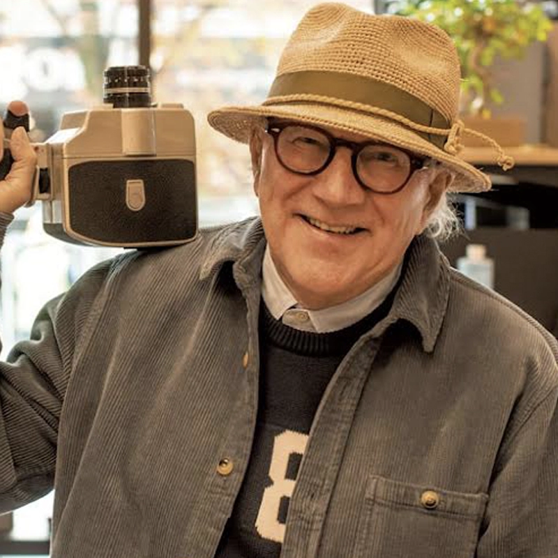 Allan Nicholls, an older man with glasses and a straw hat, smiles while holding a vintage film camera on his shoulder. He wears a grey corduroy jacket over a sweater with the number 8; blurred indoor plants and shelves are in the background.