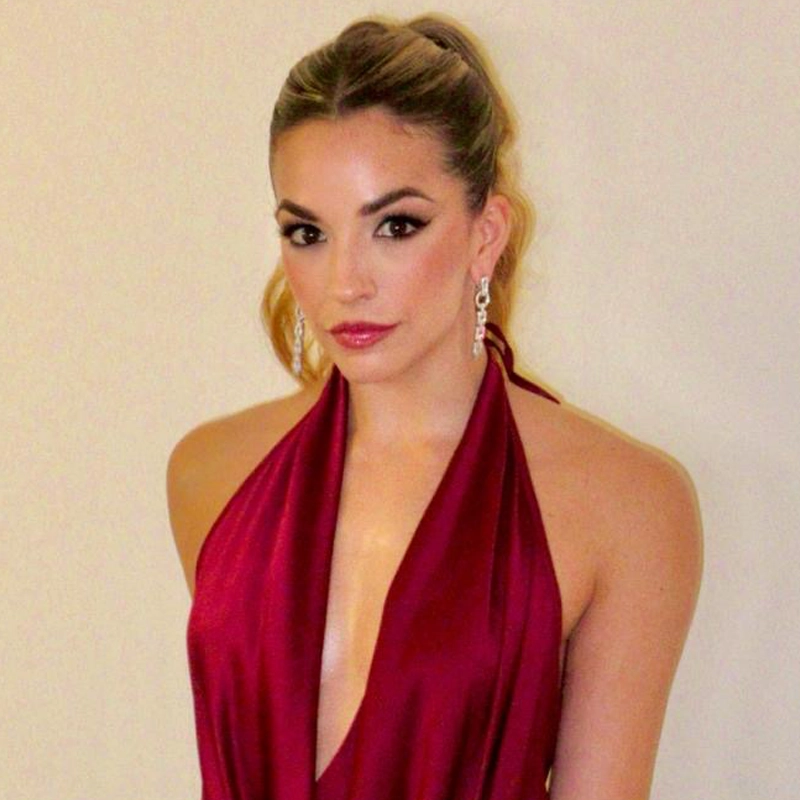 Ana Villafañe, with light skin and long, pulled-back hair, wears a deep red halter dress with a plunging neckline. Her dramatic eye makeup, glossy lips, and dangling earrings stand out as she looks directly at the camera against a light background.