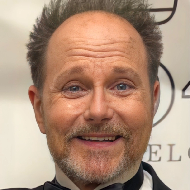 A middle-aged man with a receding hairline, tousled brown hair, and a trimmed beard smiles at the camera. Walter Willison is wearing a black tuxedo with a white shirt and bowtie, standing against a light background with partial, blurry text.