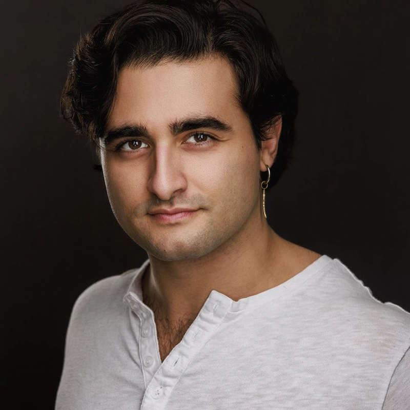 A man with wavy dark hair, fair skin, and dark eyebrows wears a white henley shirt and a gold hoop earring. He faces forward and smiles slightly against a dark background—echoing the classic charm of Giuseppe Bausilio.